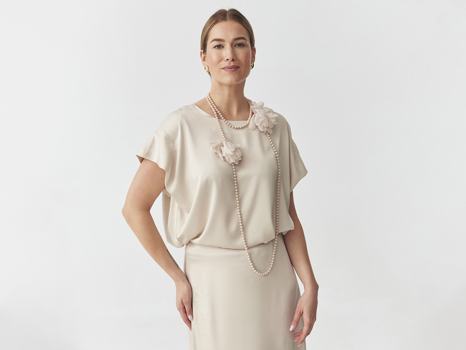 Žena v elegantním ecru outfitu s květinovou ozdobou a dlouhým perlovým náhrdelníkem.